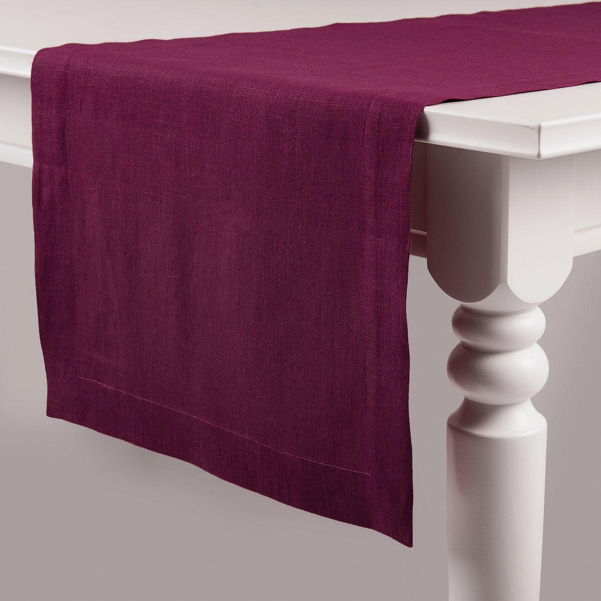 Deep Burgundy natural linen table runner over a table with white legs, showing classic deep hems and mitered corners.
